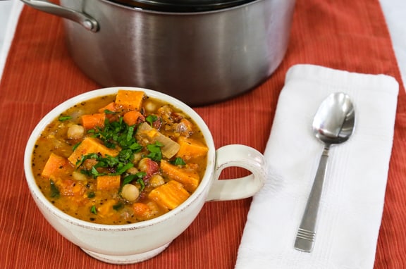 Bowl of chunky sweet potato bisque next to a stainless steel soup pot and a white napkin with a spoon