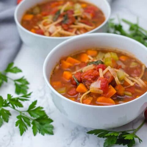 Two white bowls of Italian minestrone noodle soup garnished with parsley and cherry tomatoes