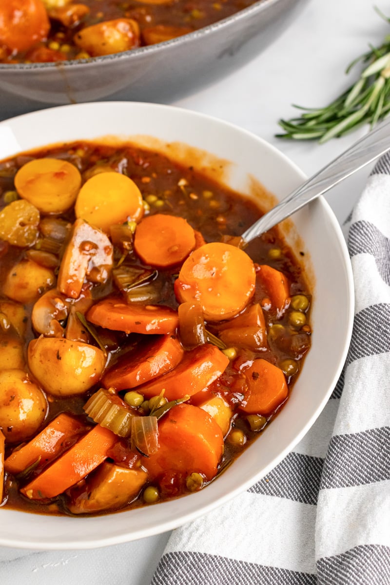 white bowl of vegan stew with potatoes, carrots, and peas next to a striped kitchen towel