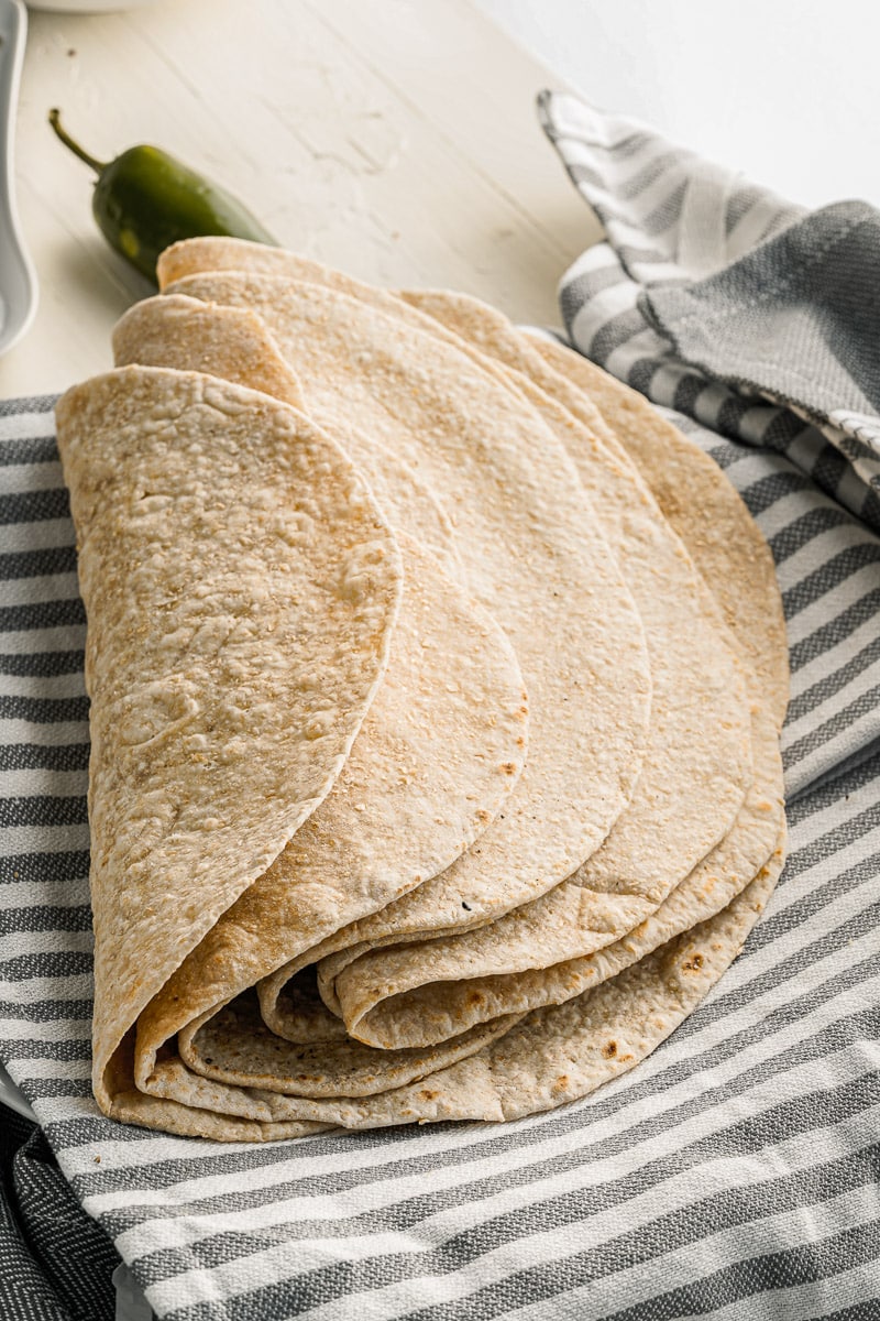 pile of homemade flour tortillas folded on a striped dish towel
