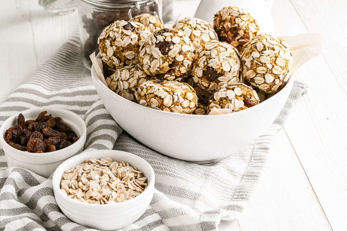 pumpkin protein balls in a white bowl next to small bowls of raisins and oats