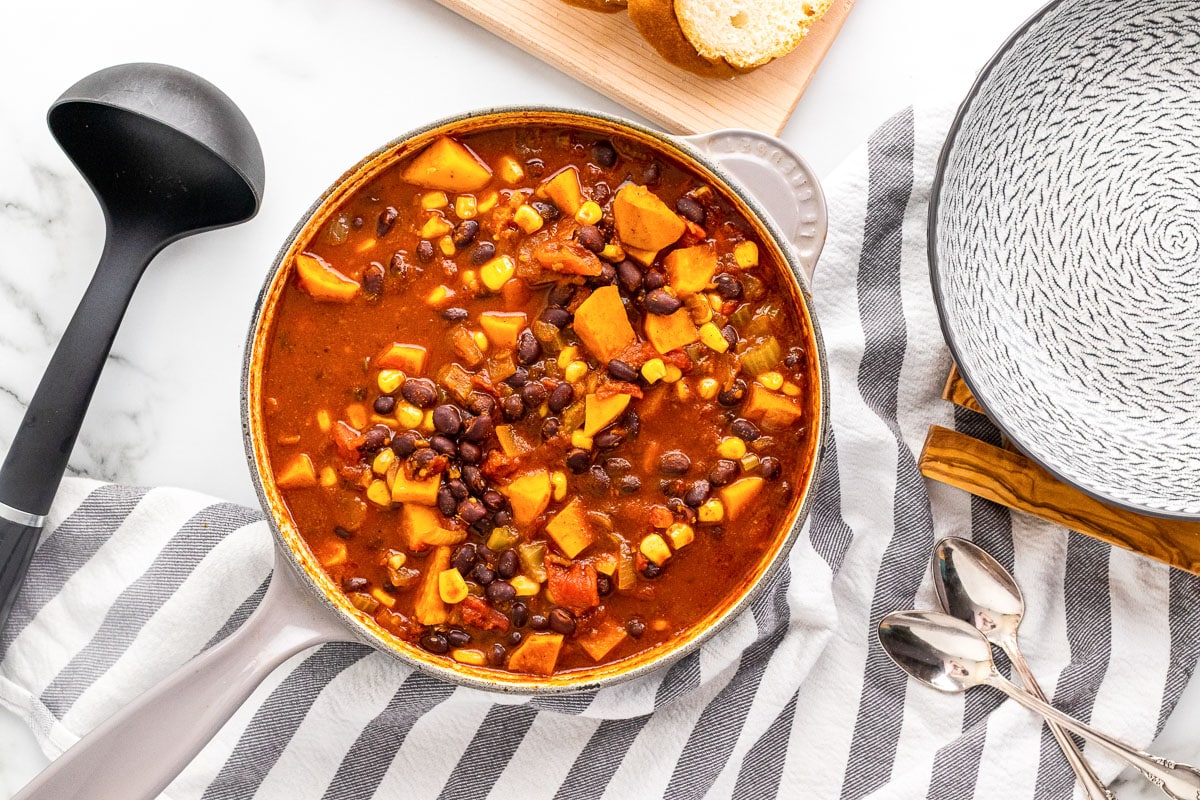 crock of sweet potato black bean chili on table with striped dishtowel, black ladle, and bread slices