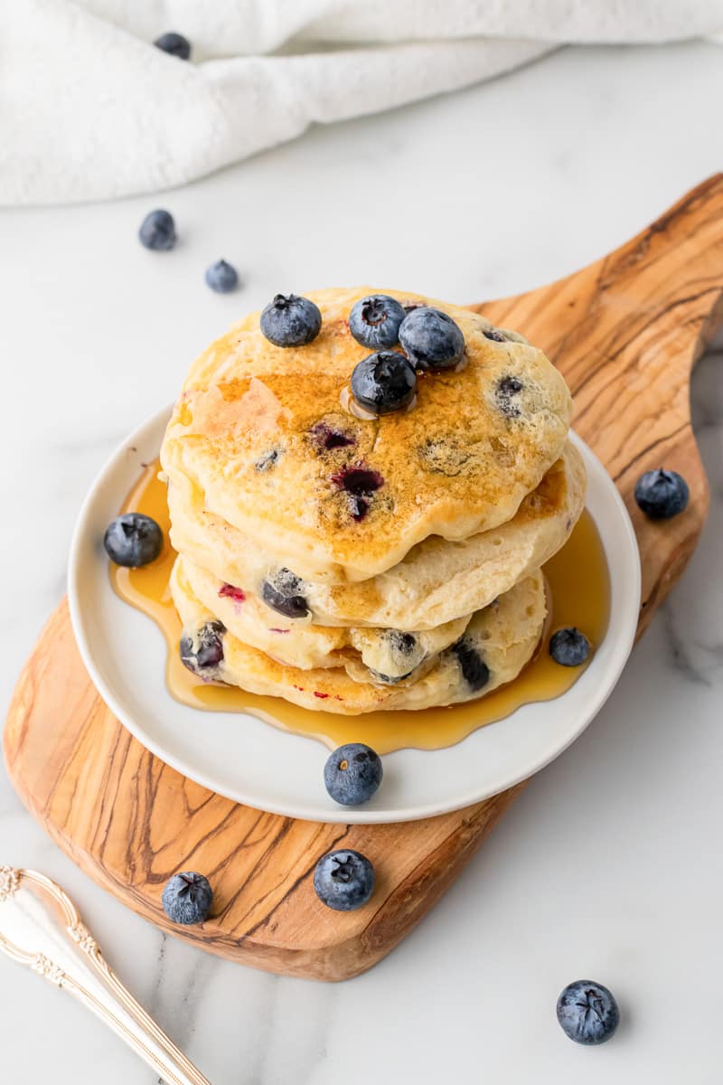 close up stack of vegan blueberry pancakes topped with blueberries and syrup on white plate on wooden board