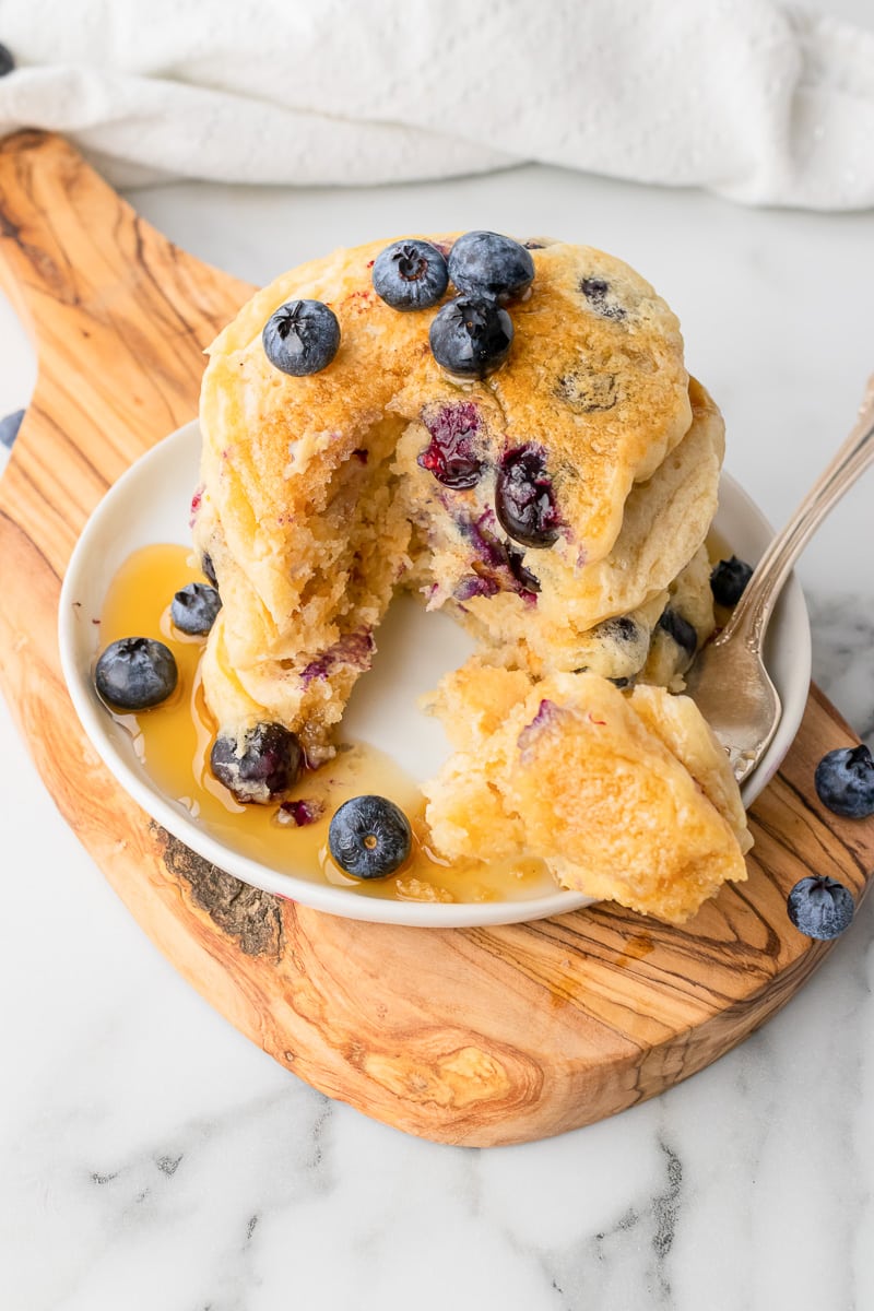 stack of vegan blueberry pancakes with a slice cut out and fork and syrup on white plate