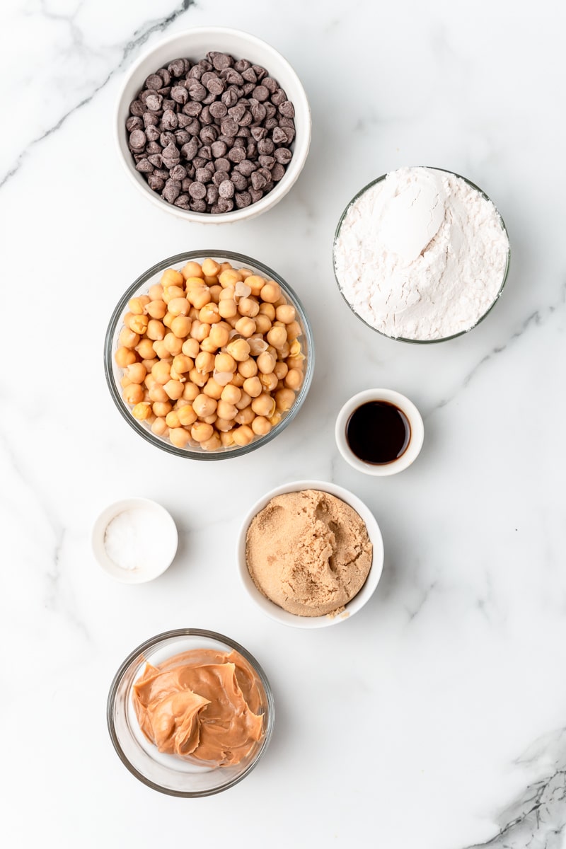 Ingredients for chickpea cookies in small bowls: chickpeas, flour, peanut butter, brown sugar, vanilla, baking soda, salt and chocolate chips