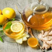 A glass teapot and cup of lemon ginger tea with fresh lemon slices, ginger root, and honey