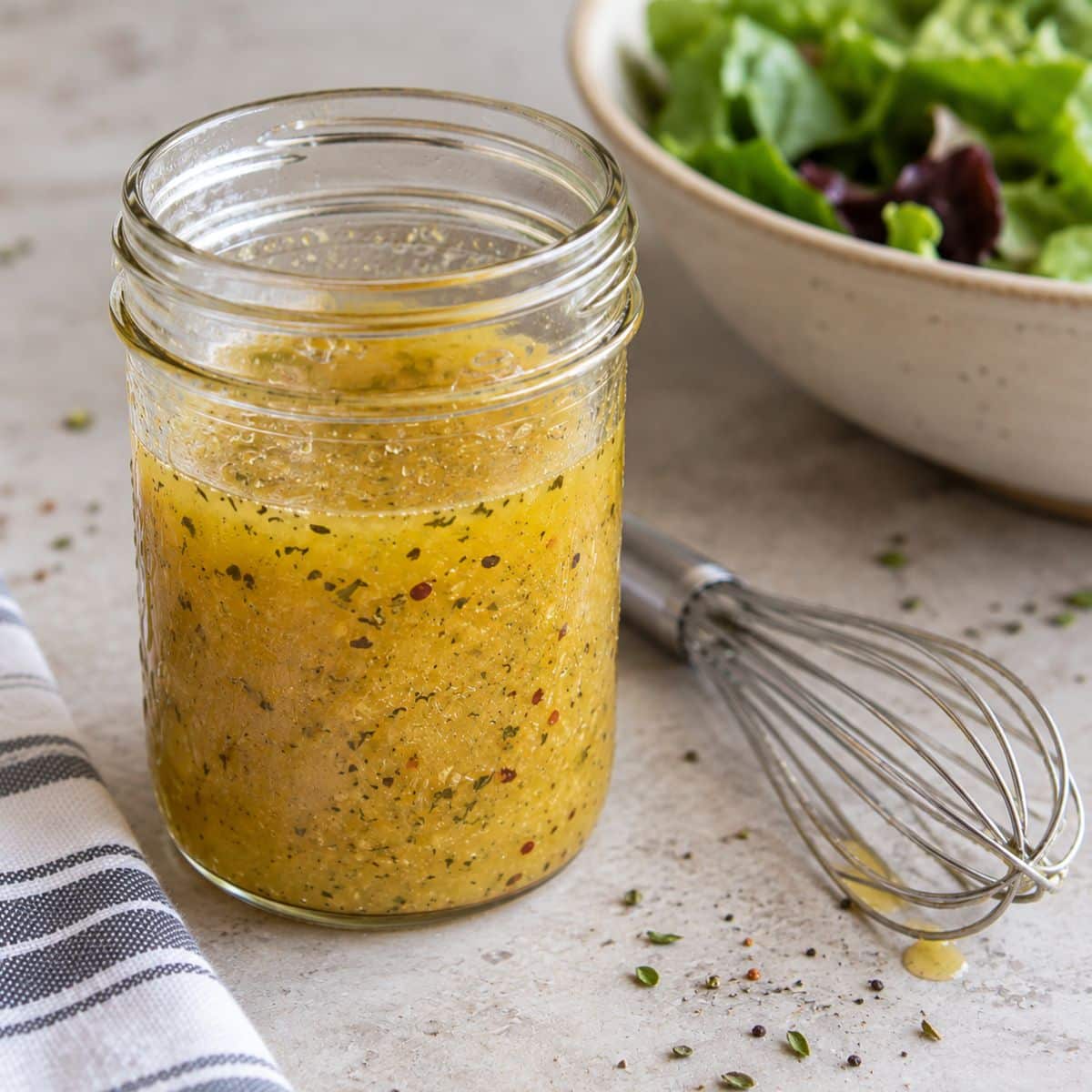 Mason jar of homemade vinaigrette dressing with herbs and red pepper flakes next to a whisk and a bowl of salad greens