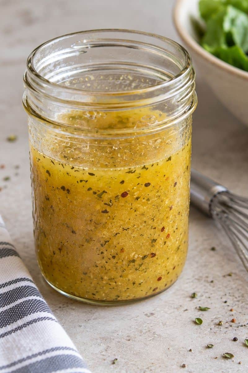 Glass jar of herby vinaigrette dressing flecked with red pepper flakes on a stone countertop