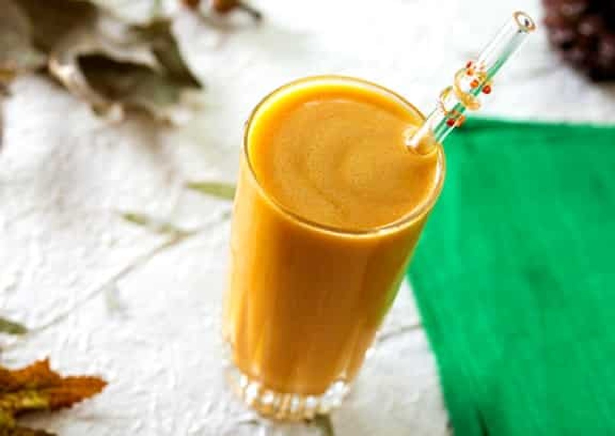A tall glass of creamy orange butternut squash smoothie with a glass straw, set on a white surface with fall leaves and a pinecone