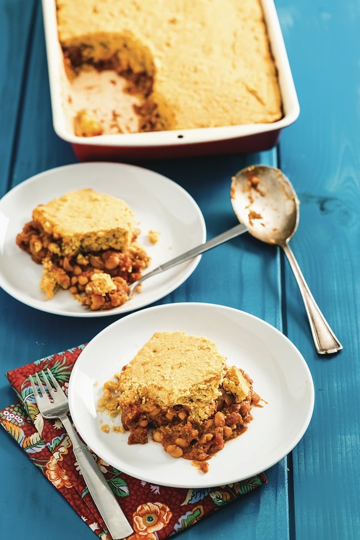 Baking dish of maple baked beans and cornbread casserole with two plated servings on a blue table