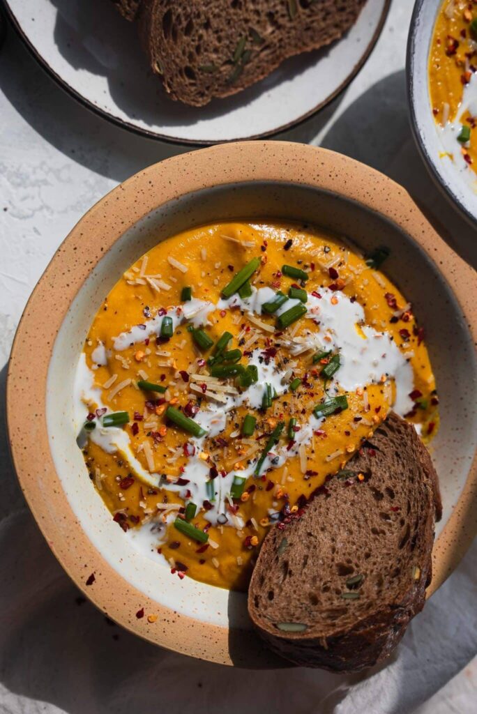 Bowl of roasted vegetable pumpkin soup topped with coconut cream, chives and chili flakes, with a slice of rye bread