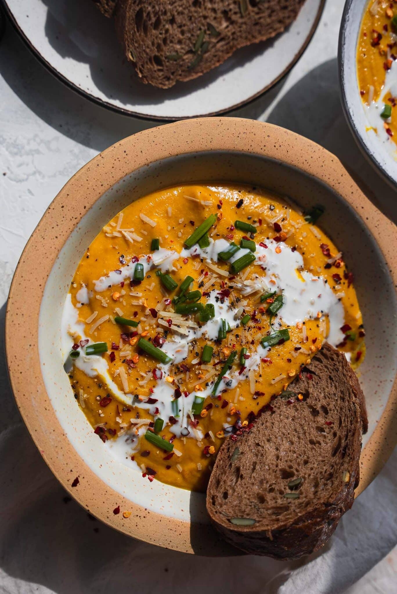 Bowl of roasted vegetable pumpkin soup topped with coconut cream, chives and chili flakes, with a slice of rye bread