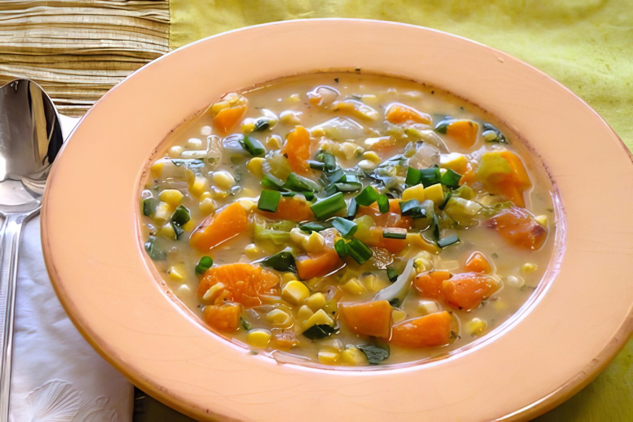 A full bowl of vegan butternut squash, sweet potato, and corn chowder with a spoon, set on a yellow linen napkin