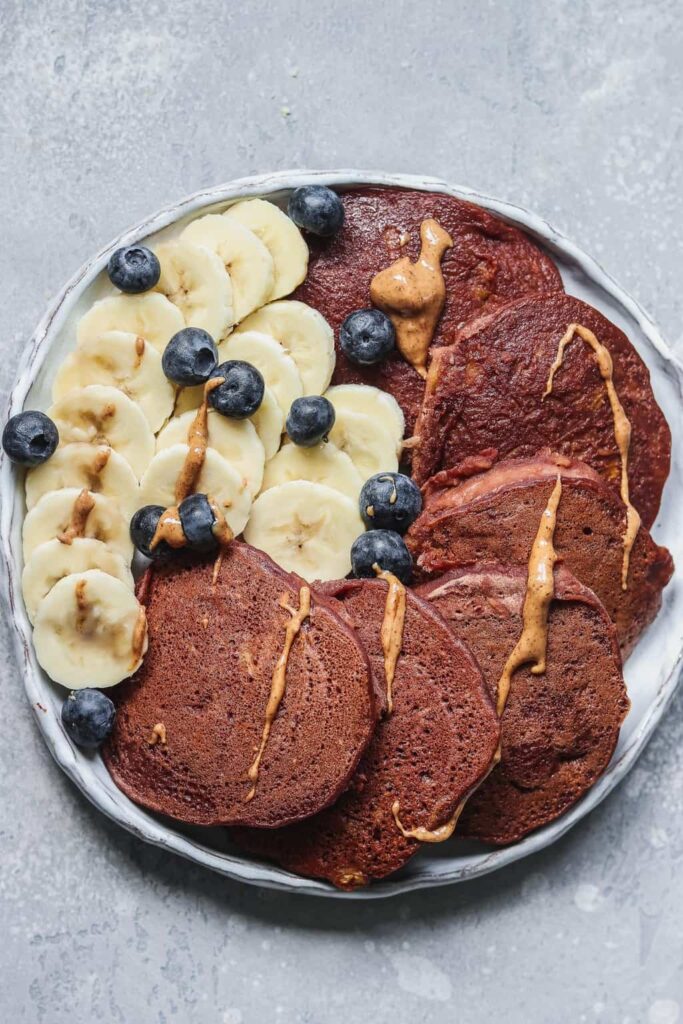 Plate of vegan chocolate pumpkin pancakes with sliced banana, blueberries and a drizzle of peanut butter