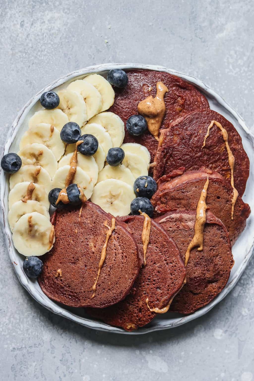 Plate of vegan chocolate pumpkin pancakes with sliced banana, blueberries and a drizzle of peanut butter