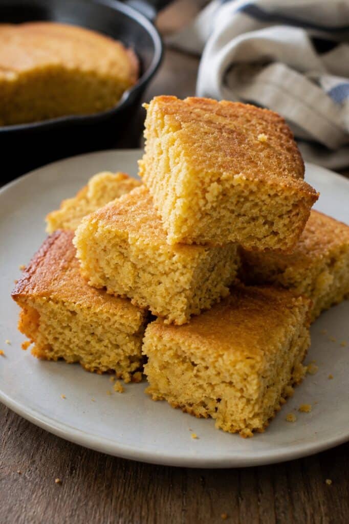 Tall stack of vegan cornbread squares on a plate with a skillet of cornbread and a striped napkin behind it