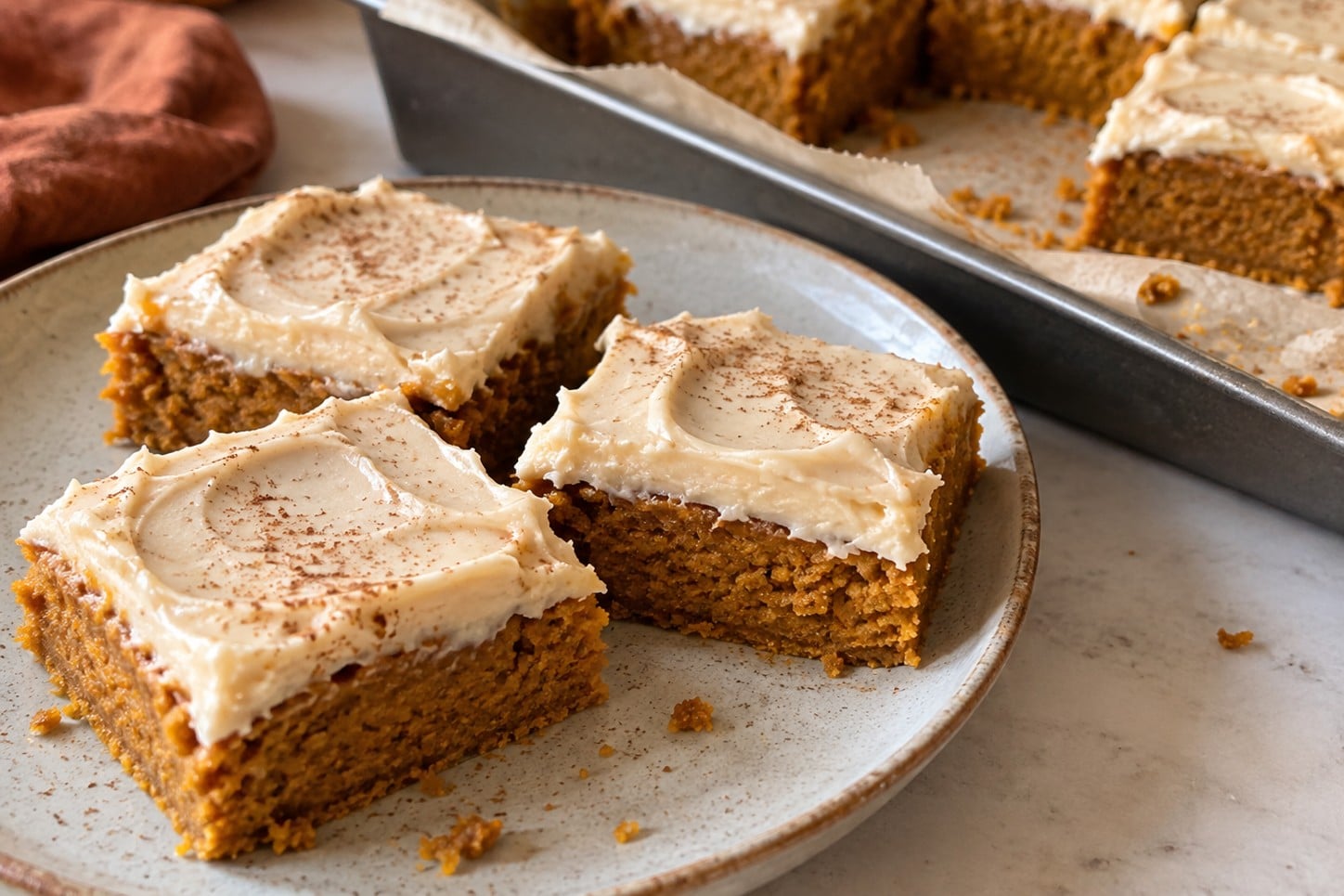 Frosted vegan pumpkin bars on a stoneware plate next to a parchment-lined baking pan of more bars