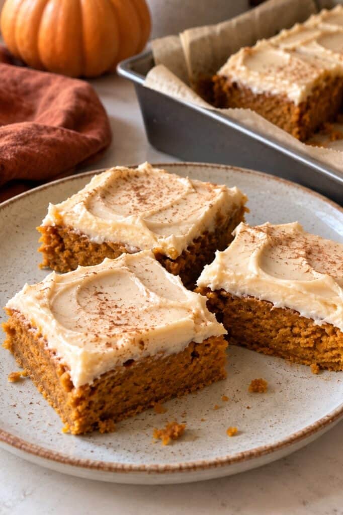 Three vegan pumpkin bars with cream cheese frosting and a dusting of cinnamon on a speckled plate, with a pumpkin and the baking pan behind