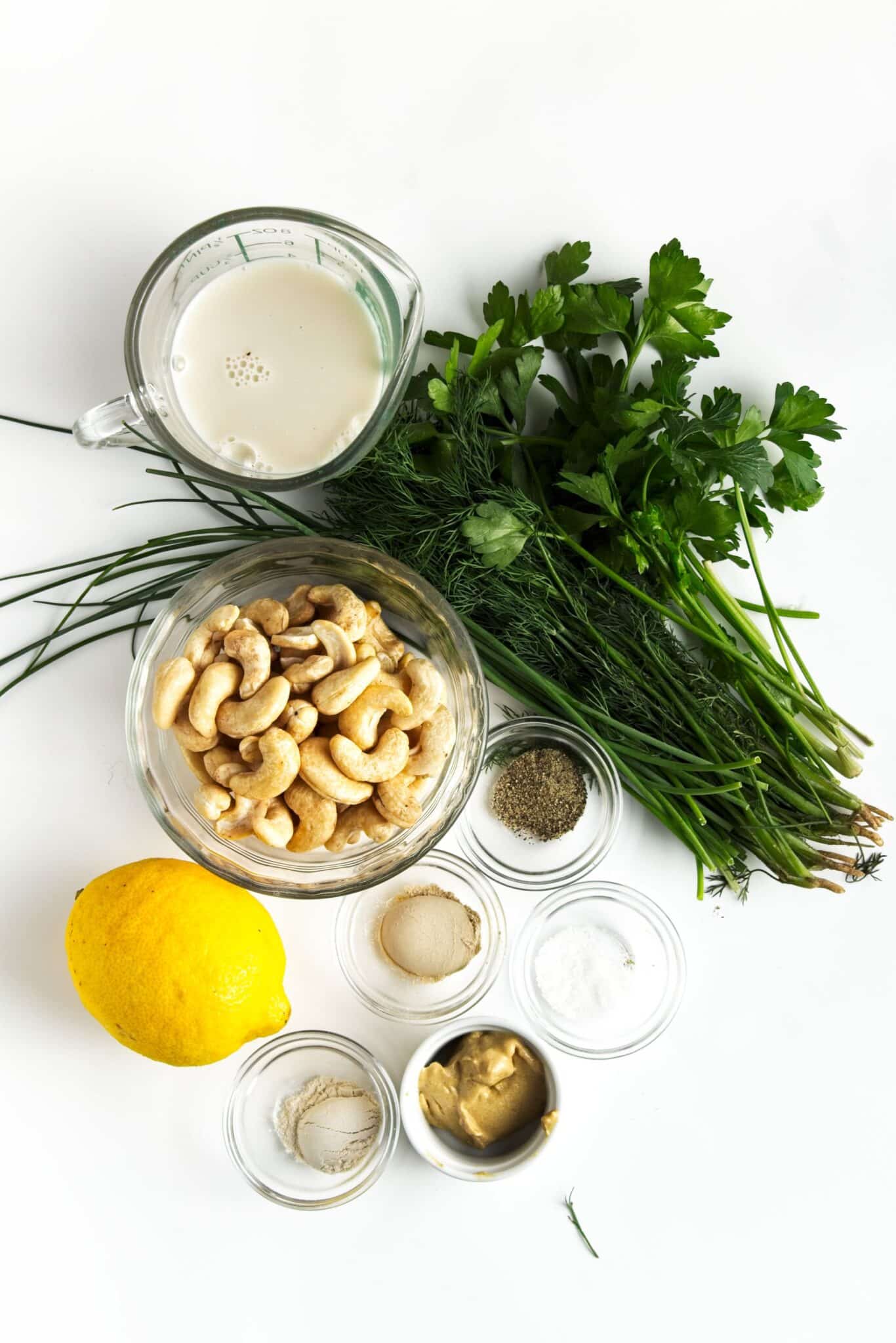 Ingredients for vegan ranch dressing including raw cashews, almond milk, lemon, garlic powder, onion powder, Dijon mustard, and fresh herbs