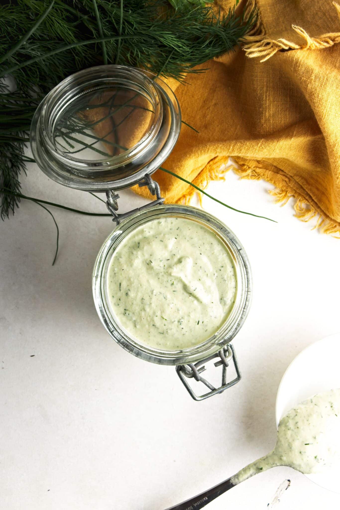 Close-up of vegan ranch dressing in an open glass jar with fresh dill and parsley in the background