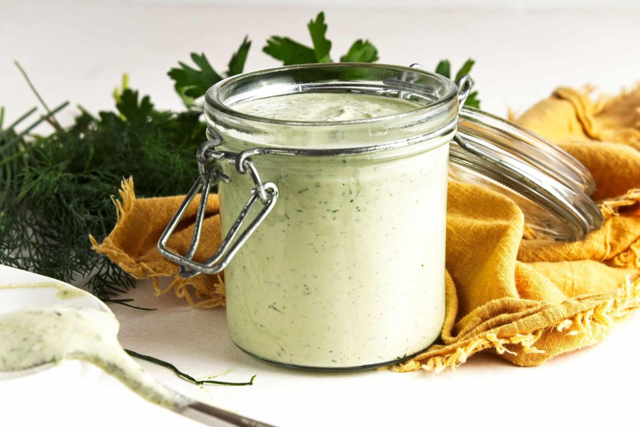 Open jar of creamy vegan ranch dressing with fresh herbs and a mustard yellow napkin