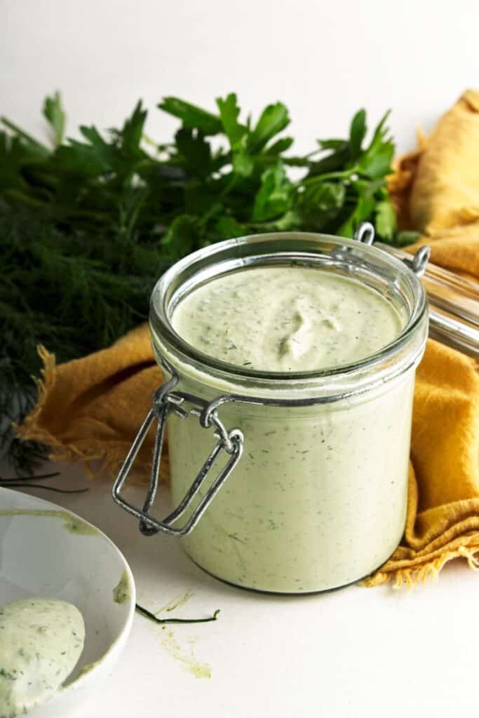 Jar of vegan ranch dressing with a spoonful in a white bowl, surrounded by fresh dill and parsley