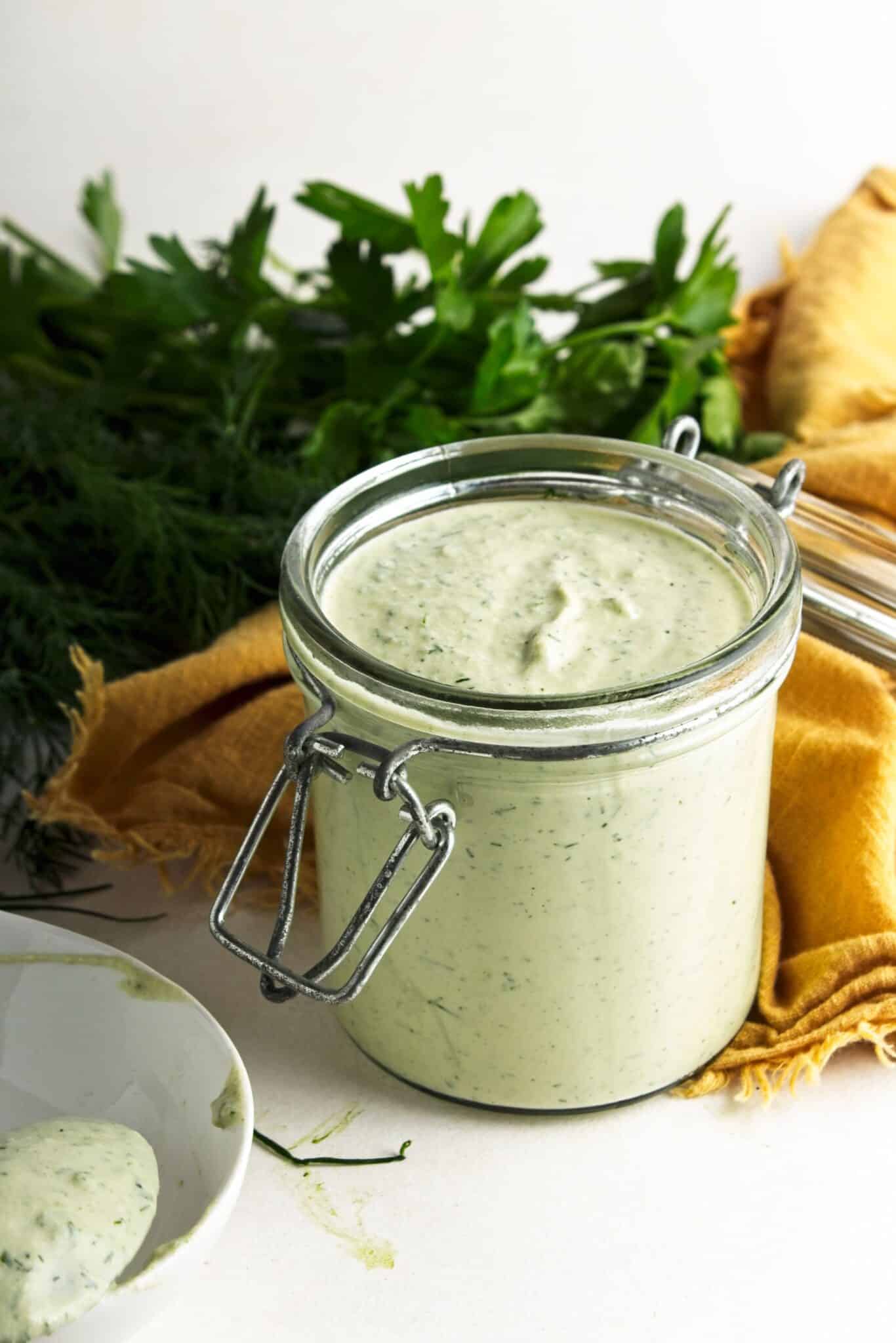 Jar of vegan ranch dressing with a spoonful in a white bowl, surrounded by fresh dill and parsley