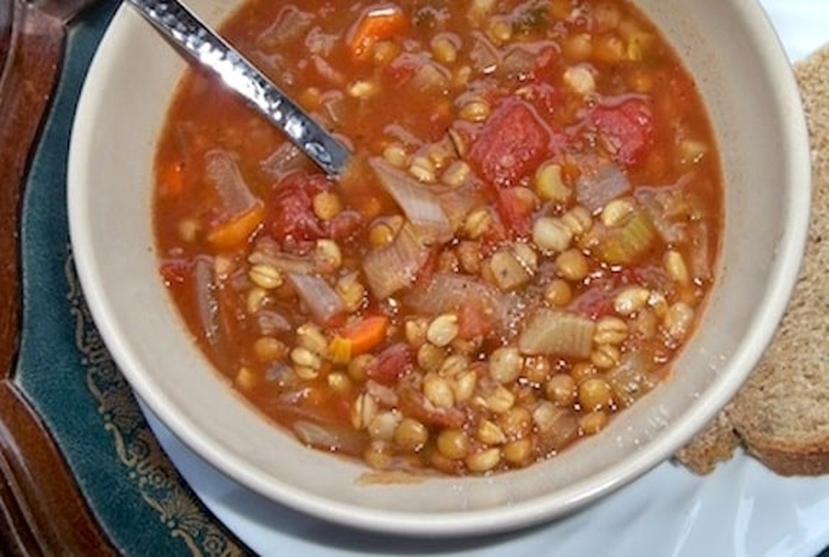 Overhead view of a bowl of vegan tomato lentil soup with barley, carrots, and celery, garnished with a spoon and served with bread