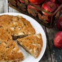 Fresh chunky applesauce cake on a white plate with a slice cut, next to a basket of red apples