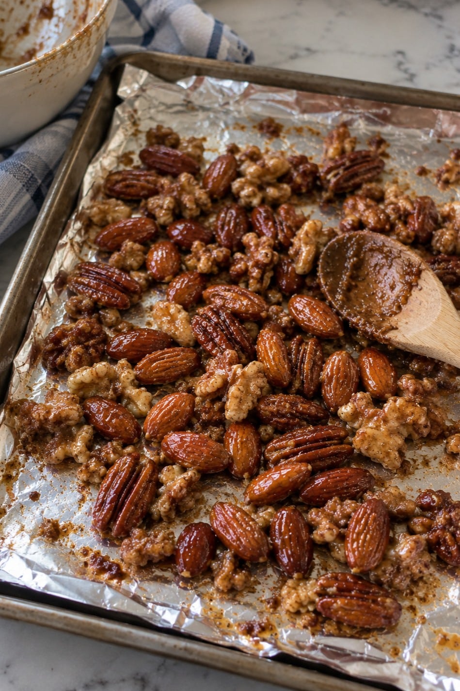 Spiced glazed nuts spread on a foil-lined baking sheet with a wooden spoon