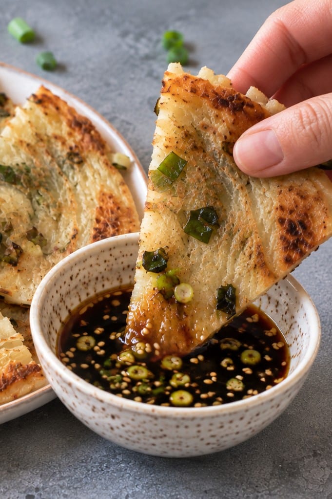 Hand dipping a wedge of vegan scallion pancake into soy dipping sauce with sesame seeds and sliced scallions